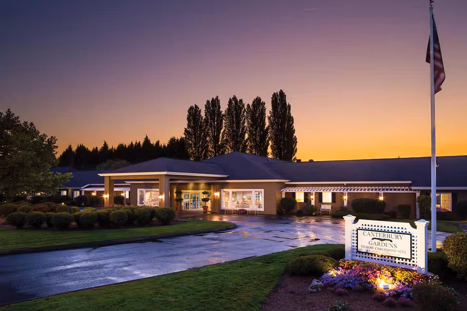 Exterior view of Canterbury Gardens Memory Care Community building at dusk with illuminated entrance, manicured bushes, and a sign in the foreground surrounded by flowers. Tall trees and a flagpole are visible in the background under a colorful sunset sky.