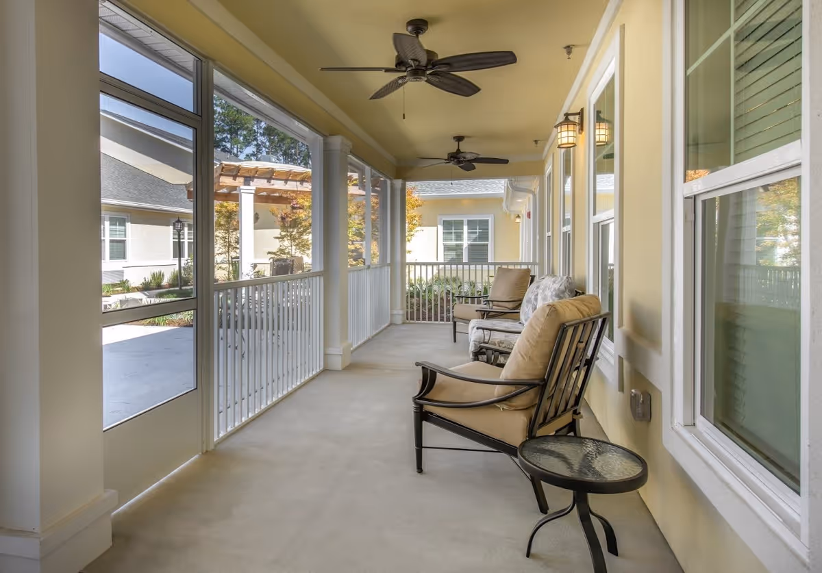 A screened-in porch area with beige walls and ceiling fans. The porch has cushioned metal chairs and a small round glass-top table. Outside the porch, there is a view of a courtyard with plants and a pergola structure.