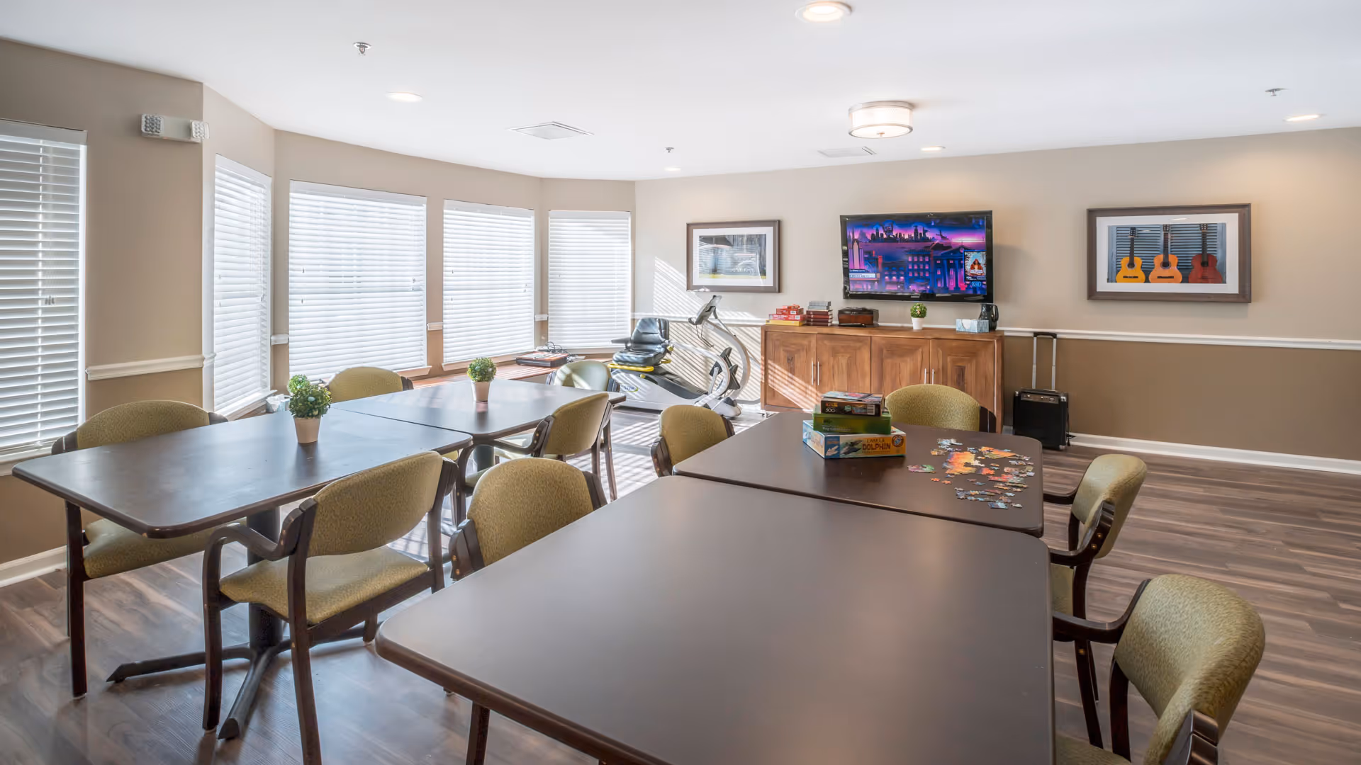 A bright activity room with several tables and chairs arranged for group activities. There are two potted plants on one table, and a partially completed jigsaw puzzle with puzzle boxes on another table. The room has large windows with blinds letting in natural light. Against the far wall, there is a wooden cabinet with a flat-screen TV mounted above it, and framed pictures on the wall. An exercise bike is positioned near the windows.