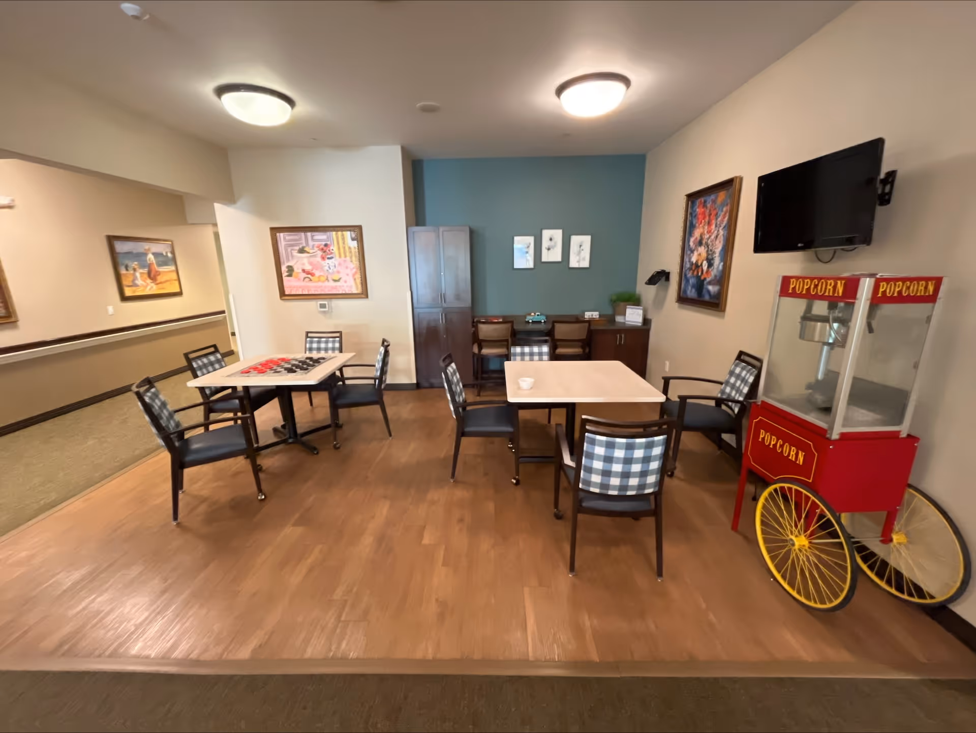 A common area with two square tables surrounded by chairs with checkered cushions. On the right side, there is a red popcorn machine with yellow wheels and a wall-mounted TV above it. The walls are decorated with framed paintings and three small artworks above a cabinet with two chairs. The floor is wooden, and the ceiling has two round light fixtures.