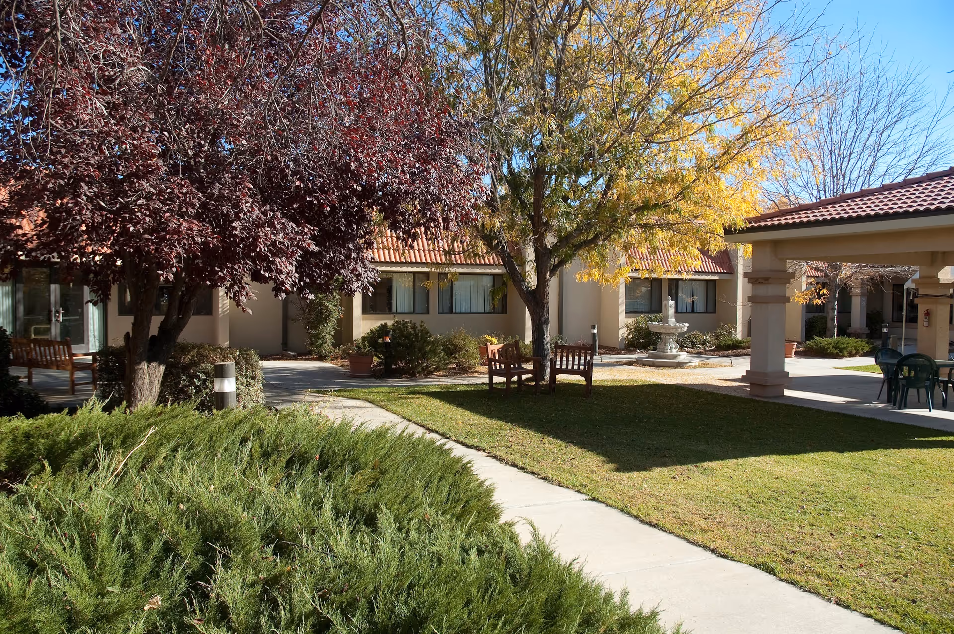 Outdoor courtyard area of a senior living facility with a paved walkway, green grass, bushes, and trees with autumn foliage. There are wooden benches under the trees and a covered patio area with chairs and tables. The building has beige walls and a red-tiled roof.