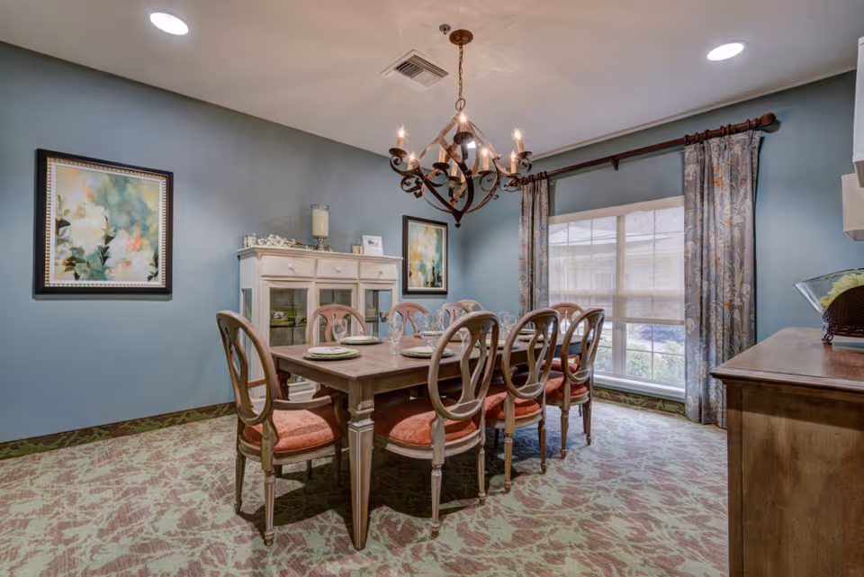 Elegant dining room with a wooden table set for eight, upholstered chairs, chandelier, sideboard, and a large window with curtains.