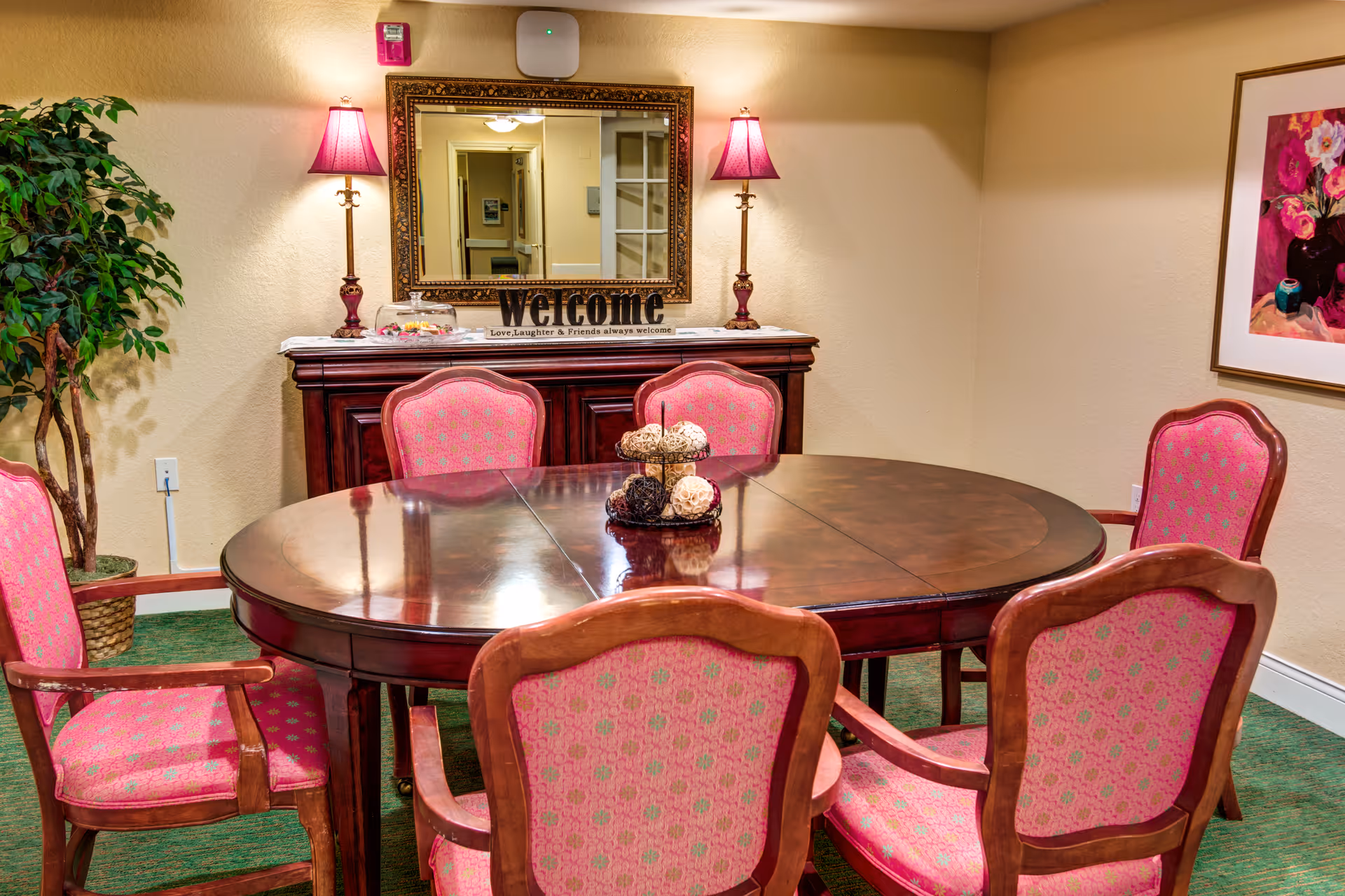Round wooden dining table surrounded by six pink upholstered chairs in a warmly lit room with a sideboard, mirror and lamps.