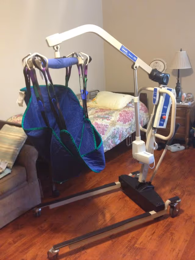 A bedroom with a floral bedspread on the bed, a beige couch with a pillow, a wooden nightstand with a lamp and some items, and a patient lift device with a blue sling in the foreground on a wooden floor.