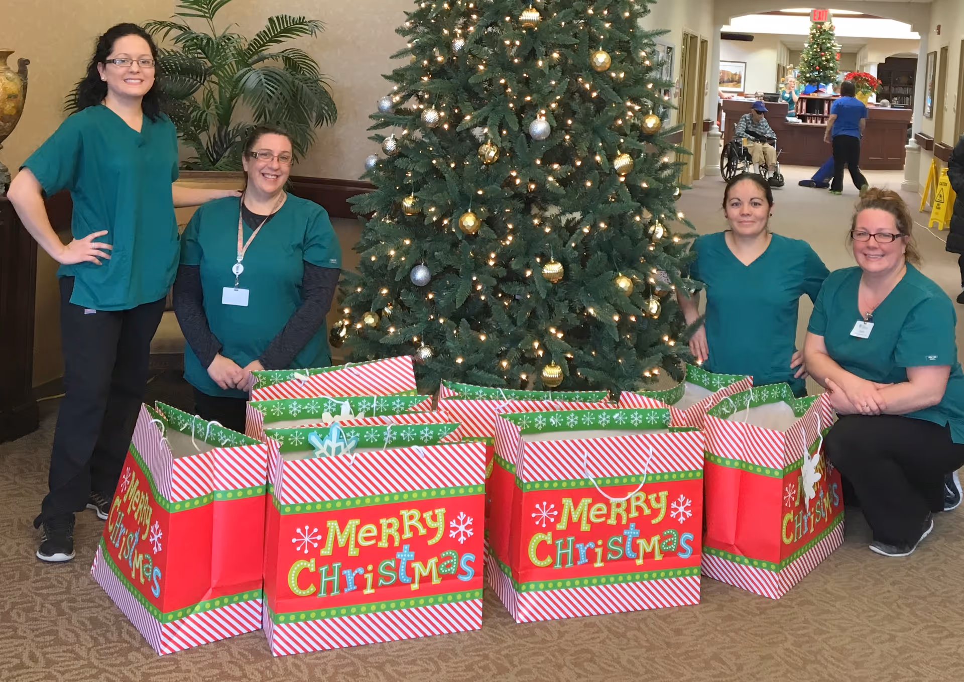 Four women in green scrubs posing around a decorated Christmas tree with several large red and green gift bags that say 'Merry Christmas' in a hallway of a senior living facility. In the background, a person in a wheelchair and other staff are visible near a reception desk.