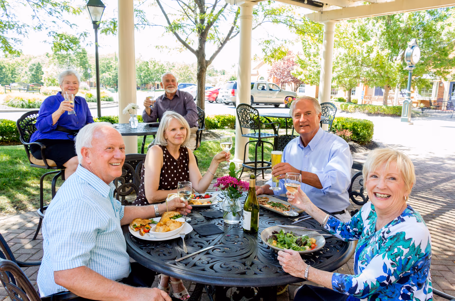 A group of six elderly people sitting outdoors at a round metal table enjoying a meal and drinks. They are smiling and raising their glasses in a toast. The setting is a sunny patio area with trees, shrubs, and parked cars visible in the background.