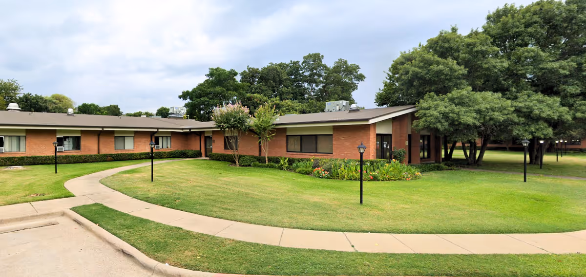 One-story brick building with a curved sidewalk, lamp posts, manicured lawn and trees under a cloudy sky.