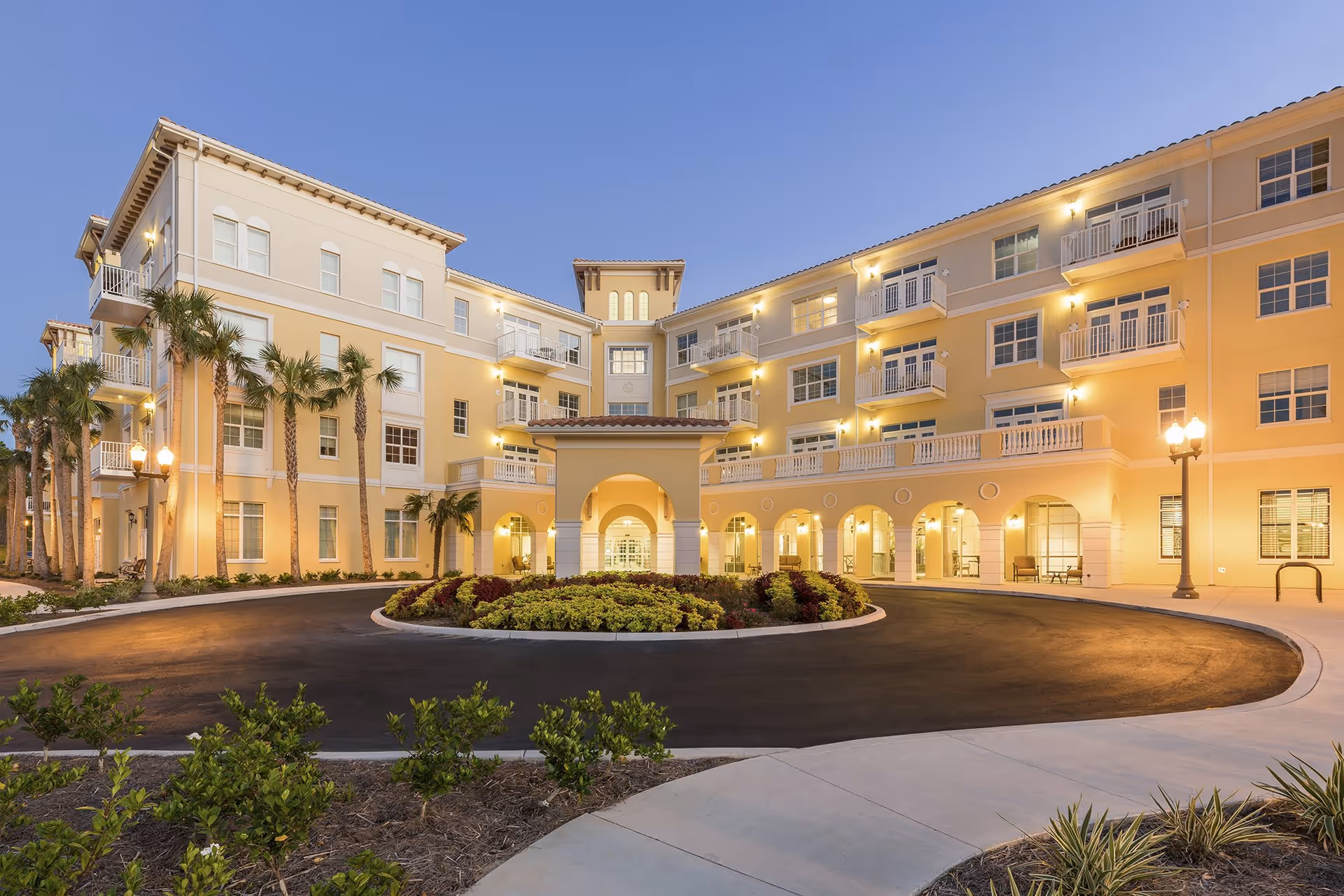 Exterior view of a large, multi-story senior living facility building at dusk with warm lighting, palm trees, a circular driveway with landscaped center, and balconies on the upper floors.
