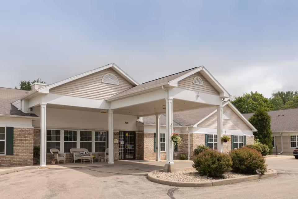 Front entrance of a single-story brick-and-siding senior living building with a covered porte-cochere, benches, and landscaped planters.