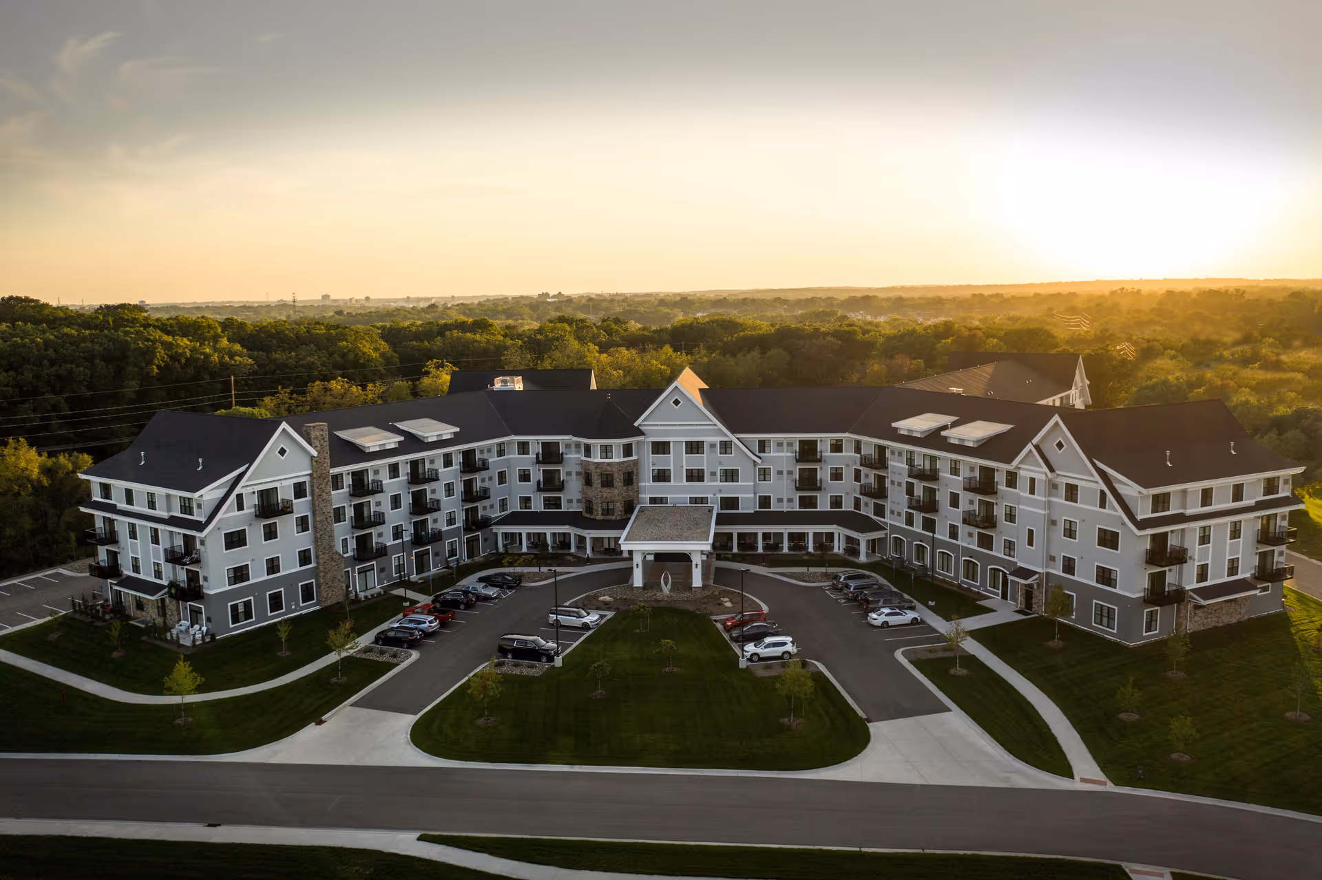 Aerial view of a large senior living facility named Grand Living At Indian Creek during sunset, surrounded by trees and greenery with a parking area in front.