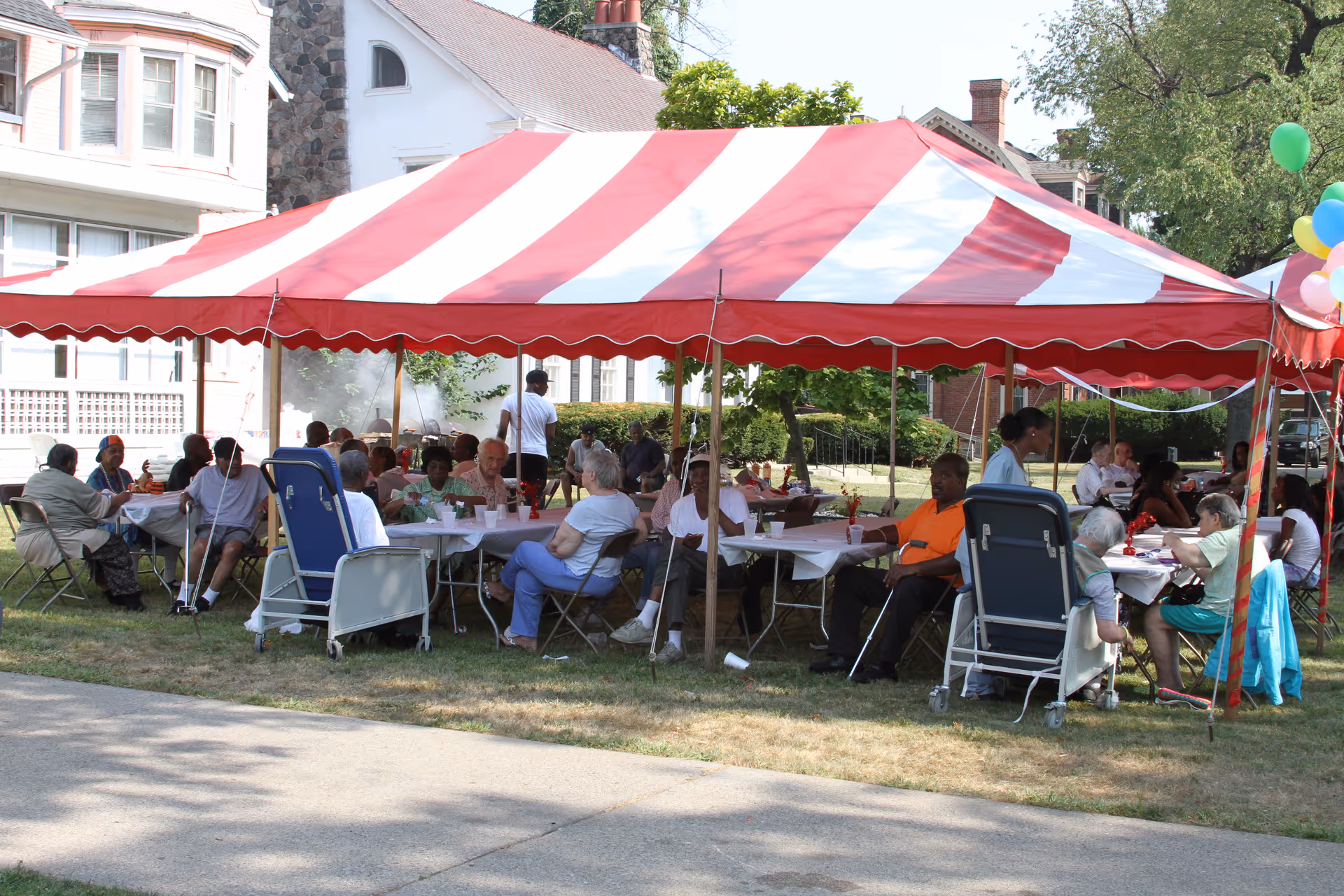 A red-and-white striped canopy tent on a lawn with elderly residents seated at tables for an outdoor gathering.