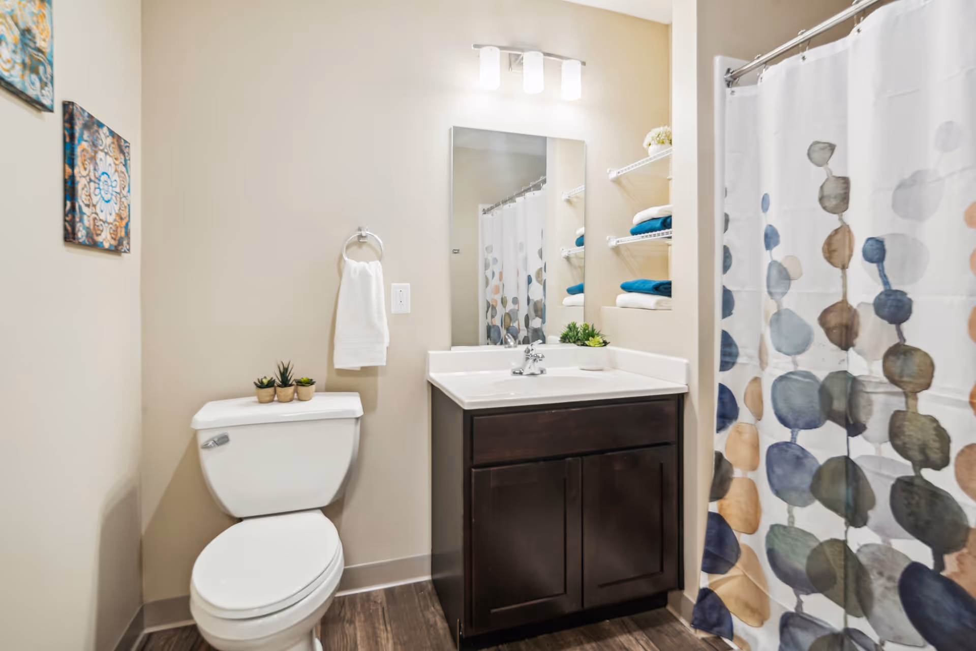 A clean and modern bathroom featuring a white toilet with small potted plants on the tank, a dark wood vanity with a white countertop and sink, a rectangular mirror above the sink, a towel ring with a white towel, built-in shelves with neatly folded towels, and a shower with a colorful patterned curtain.