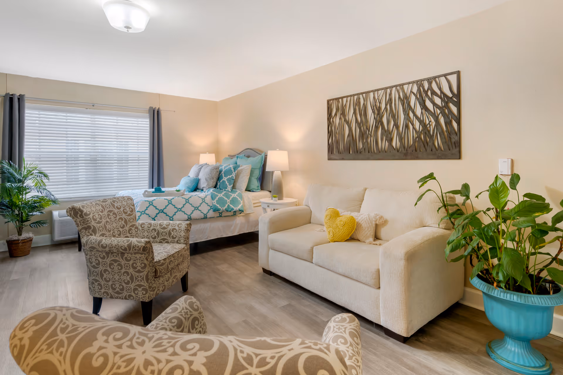 Bright studio-style living area with a bed, sofa, patterned armchairs, potted plants, and wall art.