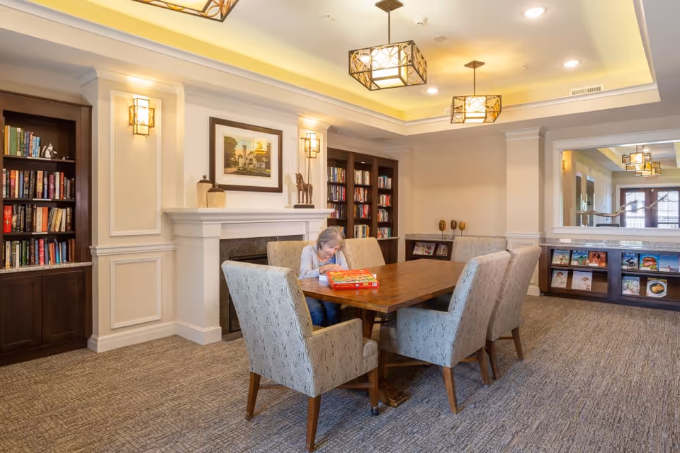 An elderly woman seated at a wooden table in a well-lit communal living room with bookshelves, a fireplace, and upholstered chairs.
