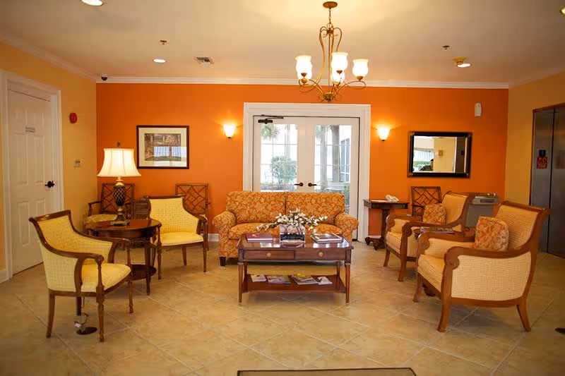 A cozy sitting area with a floral patterned sofa, four cushioned armchairs, a wooden coffee table with magazines, and a side table with a lamp. The walls are painted orange and cream, with framed artwork and a mirror. Double glass doors lead outside, and an elevator is visible on the right side.
