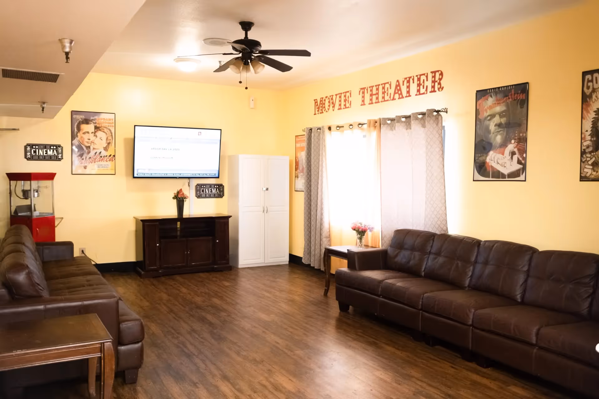 A cozy movie theater room with two brown leather sofas facing a wall-mounted flat screen TV. The walls are painted yellow and decorated with vintage movie posters and signs that say 'CINEMA' and 'MOVIE THEATER'. There is a popcorn machine on the left side and a ceiling fan with lights above. The floor is wooden, and there are curtains on the window letting in natural light.