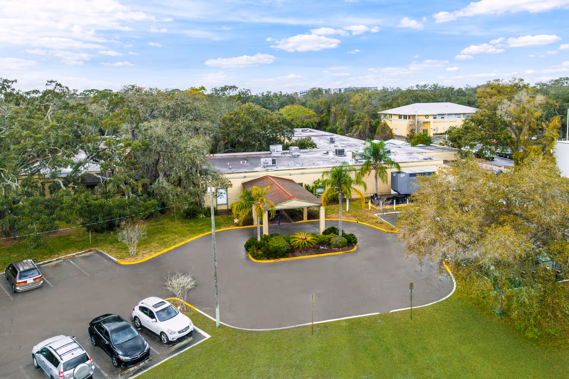 Aerial view of Fairway Oaks Center's entrance showing a circular driveway with palm trees, parked cars, and surrounding low-rise buildings and trees.