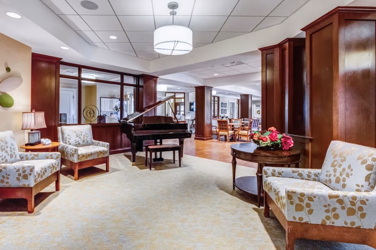 Interior view of a senior living facility lounge area featuring patterned armchairs, a round wooden table with a floral arrangement, a grand piano with a bench, and a dining area with wooden chairs and tables in the background. The space has warm lighting, wood paneling, and a carpeted floor.