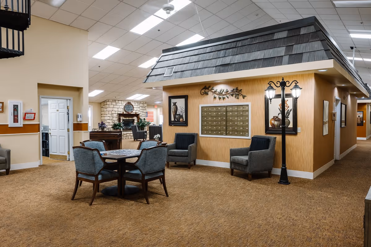 Interior common area of a senior living facility featuring a small seating area with four upholstered chairs around a round table, additional armchairs against a wall with decorative artwork and a mailbox unit, a decorative street lamp, and a stone fireplace in the background.