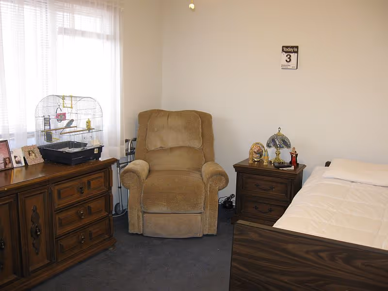 A cozy senior living bedroom featuring a beige armchair, a wooden dresser with a birdcage and framed photos on top, a wooden nightstand with decorative items and a lamp, and a bed with white bedding. A calendar is mounted on the wall above the nightstand.