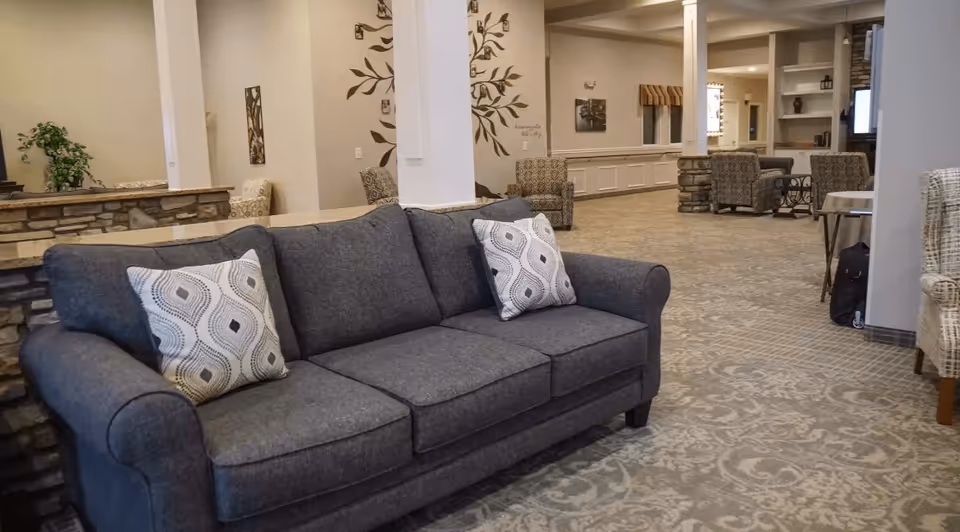 A spacious and well-lit living room area in a senior living facility featuring a gray sofa with patterned cushions, several armchairs, a stone accent wall, decorative wall art, and a patterned carpet. The room has a warm and inviting atmosphere with neutral tones and comfortable seating arrangements.