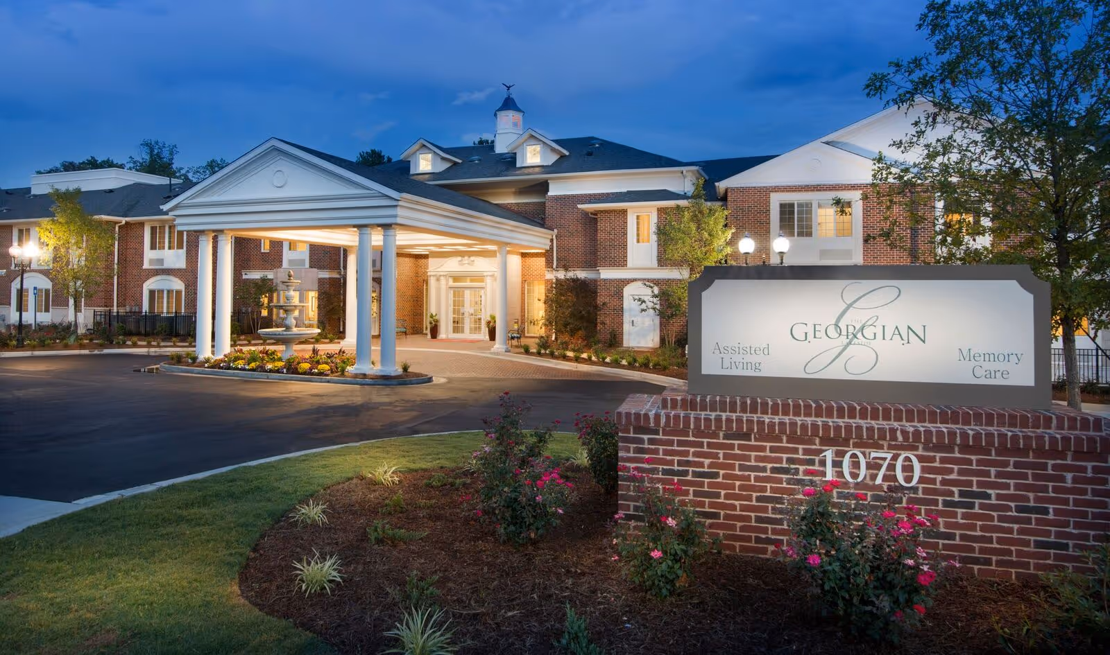 Front entrance of The Georgian assisted living facility at dusk, showing a covered porte-cochère, fountain, landscaped grounds and an illuminated sign reading "Georgian".