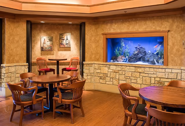 Interior view of a senior living facility common area with wooden round tables and chairs arranged on a wooden floor. There is a built-in aquarium with fish and aquatic plants embedded in a stone wall. The walls are painted in warm tones with two framed pictures hanging above a small table with two chairs in a cozy nook.