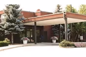 Exterior view of a building entrance with a covered drop-off area supported by columns, surrounded by trees and plants, including two large potted plants near the entrance.