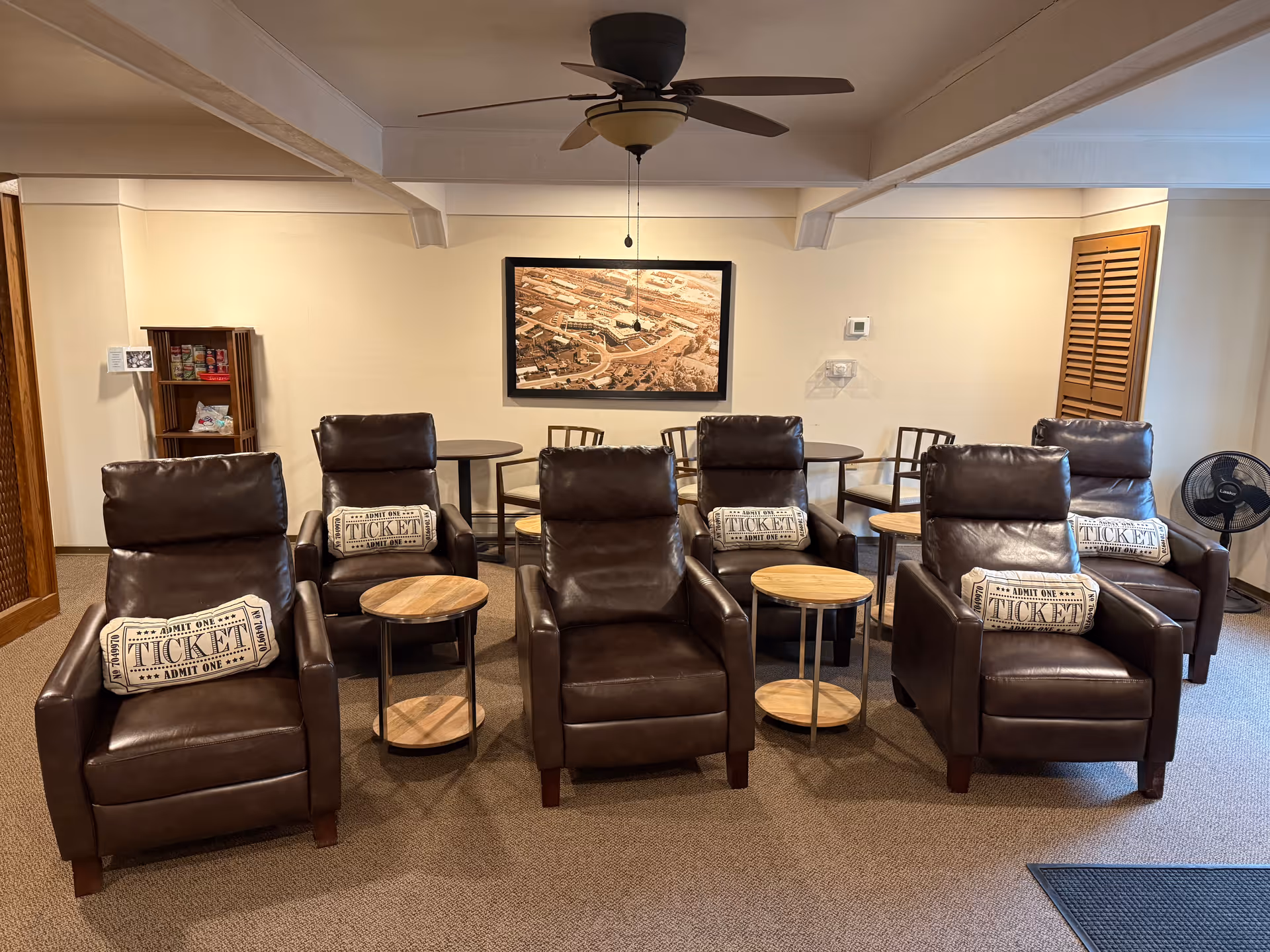 A common seating area with six brown leather recliners arranged in two rows, small round side tables, ticket-pattern pillows, a ceiling fan, and a framed aerial photo on the wall.