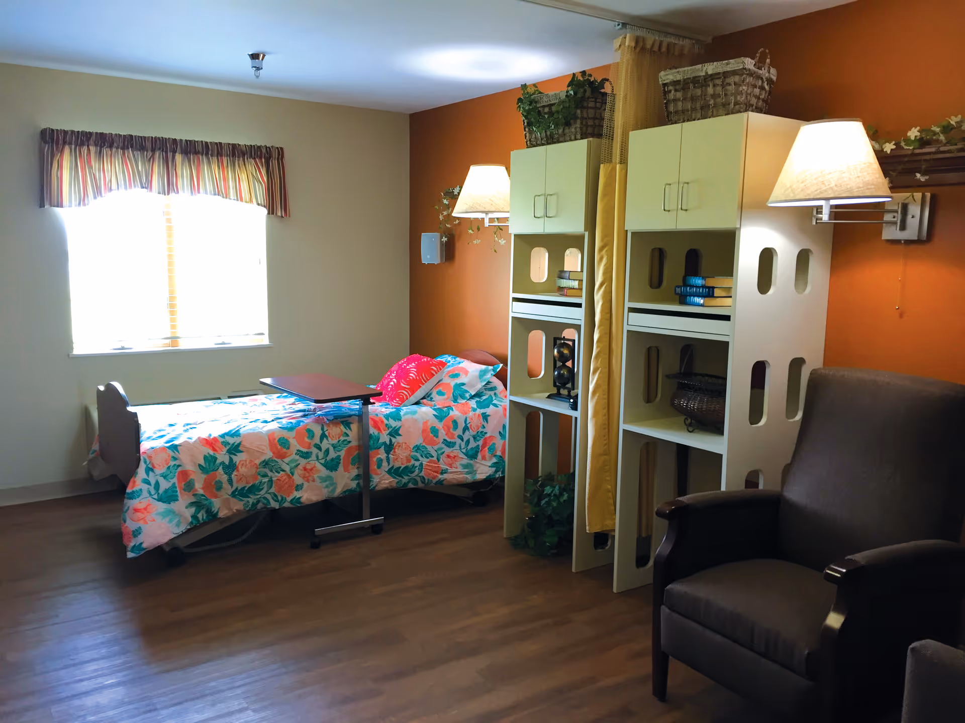 A cozy bedroom in a senior living facility with a single bed covered in a floral bedspread, a small overbed table, a window with striped valance curtains, two white shelving units with books and decorative items, wall-mounted lamps, and a comfortable armchair.