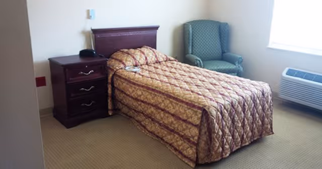 Single-occupancy bedroom with a twin bed covered by a patterned bedspread, a wooden nightstand with a phone, a green upholstered armchair, and a window air conditioner.
