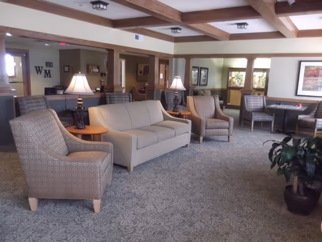 A spacious living room area with patterned carpet, a beige sofa, two patterned armchairs, two side tables with lamps, and additional seating with tables in the background. The room has exposed wooden ceiling beams and framed artwork on the walls. A potted plant is visible in the foreground.