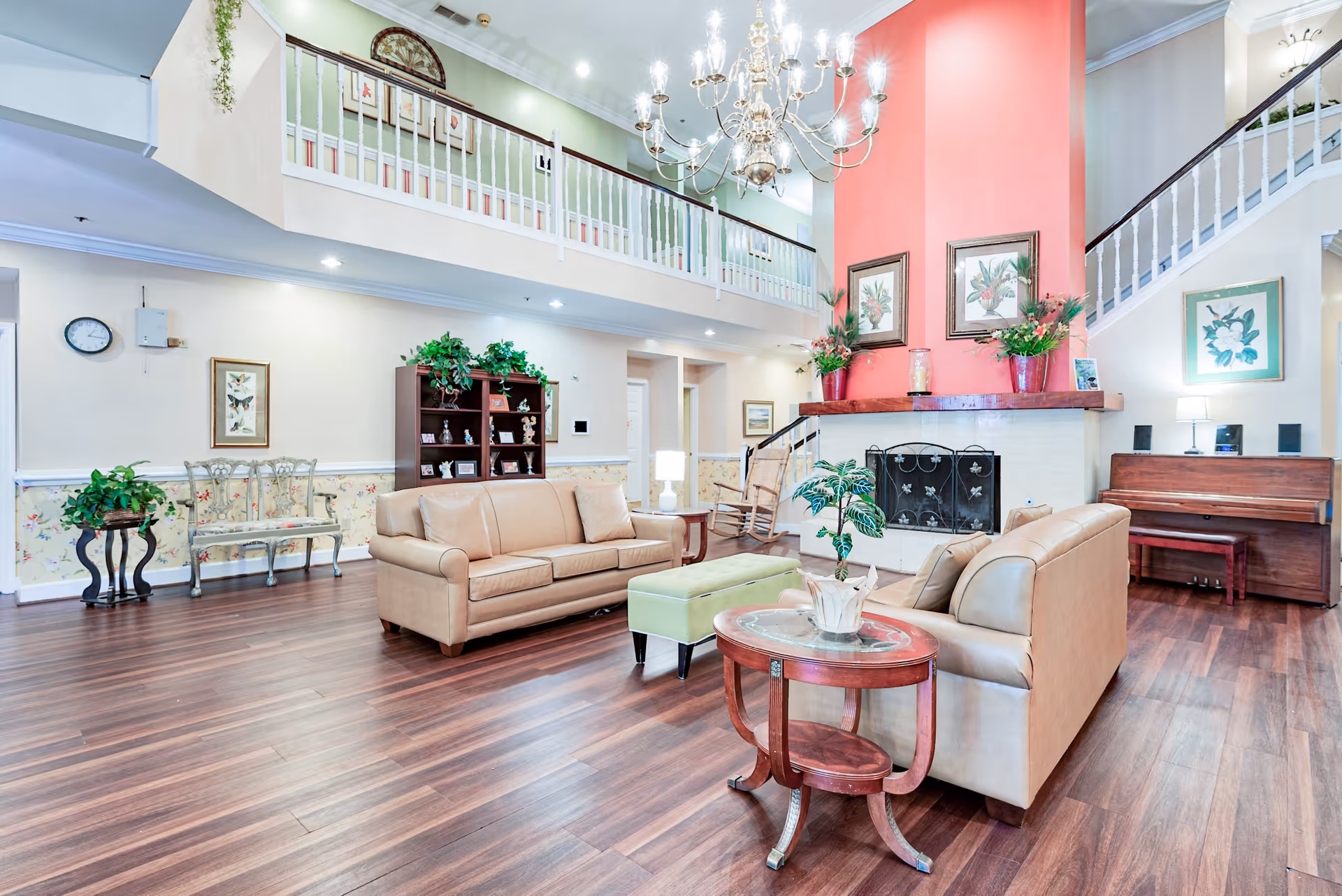 A spacious senior living facility common area with two beige leather sofas, a green ottoman, a round wooden side table with a plant, a fireplace with a red chimney, framed botanical artwork, a chandelier, a wooden piano, and a staircase with white railings leading to an upper level balcony.