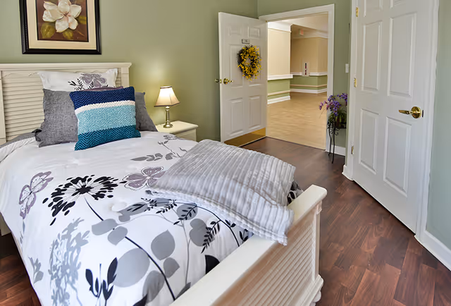 A neatly made single bed with floral patterned bedding and multiple pillows in a softly lit bedroom. A small bedside table with a lamp is next to the bed. The room has light green walls and wooden flooring. The door is open, showing a hallway with light-colored walls and flooring, decorated with a wreath on the door and a small plant stand with purple flowers.