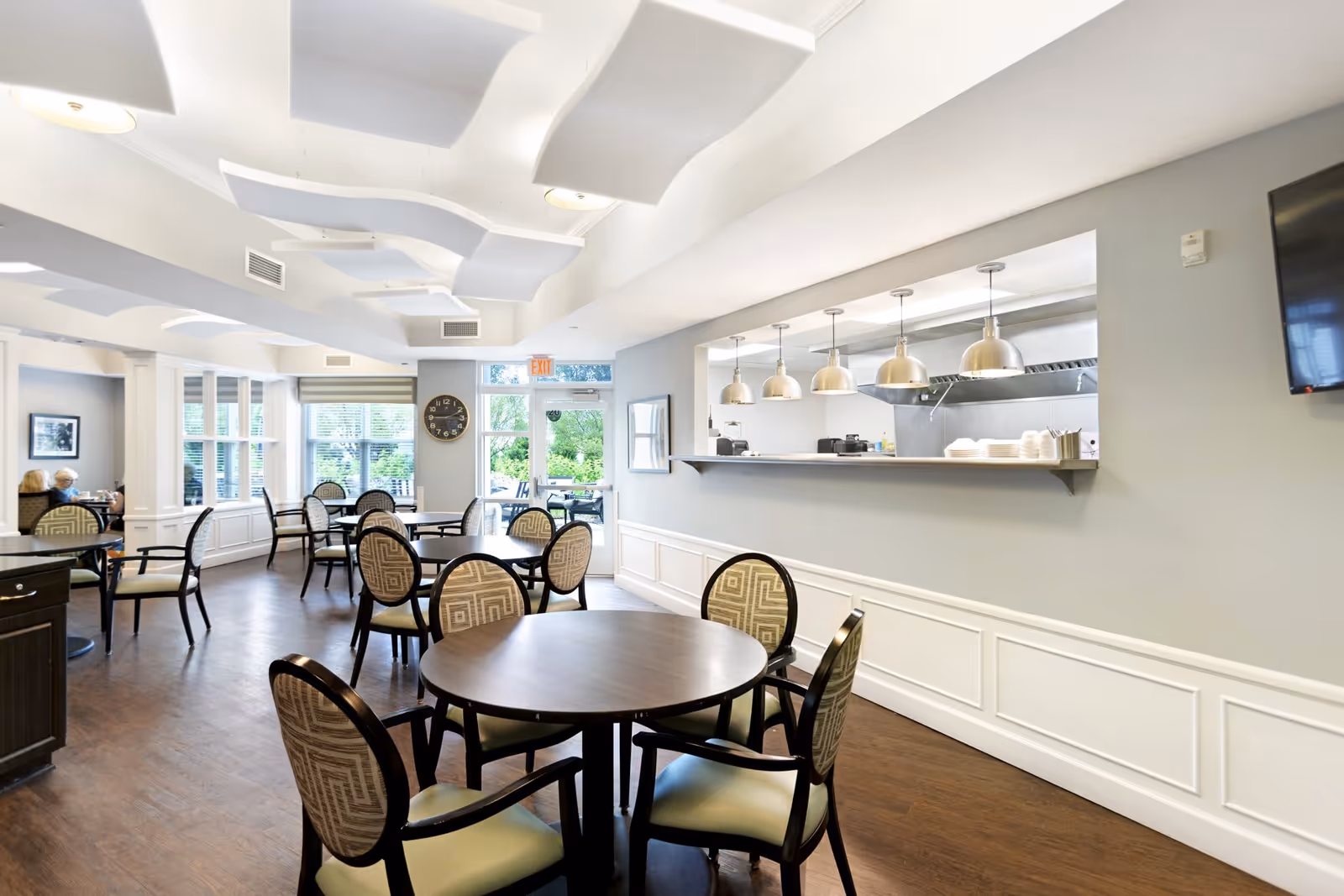 A bright and modern dining room in Olivia's Assisted Living facility featuring round tables with patterned cushioned chairs, large windows letting in natural light, a serving counter with hanging pendant lights, and a view of an outdoor patio area through a glass door.