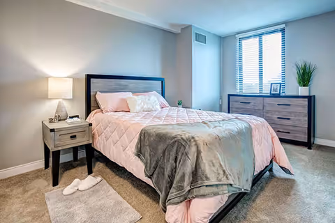 A neatly made bedroom with a bed, nightstand and lamp, dresser under a window, and soft carpeting.