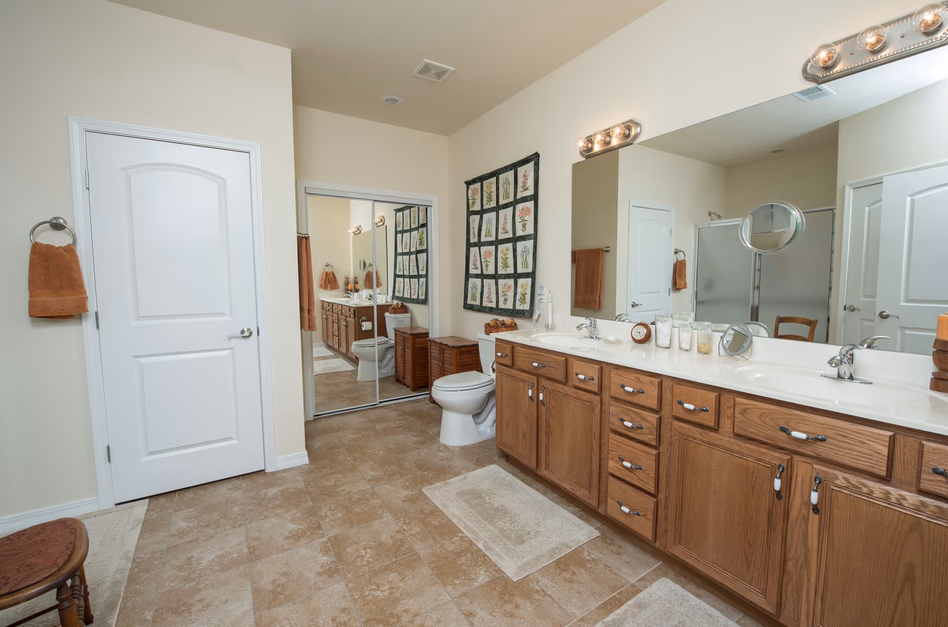A spacious bathroom with beige tiled floor, a double sink vanity with wooden cabinets, a large mirror with lights above, a toilet, a shower with frosted glass doors, a white door, and a wall hanging with floral designs. There are brown towels hanging on towel rings and a small wooden stool in the corner.