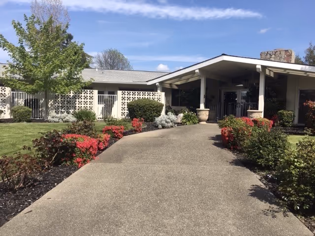 Exterior view of a single-story building with a covered entrance, surrounded by well-maintained landscaping including green grass, bushes, and red flowering plants under a blue sky with some clouds.