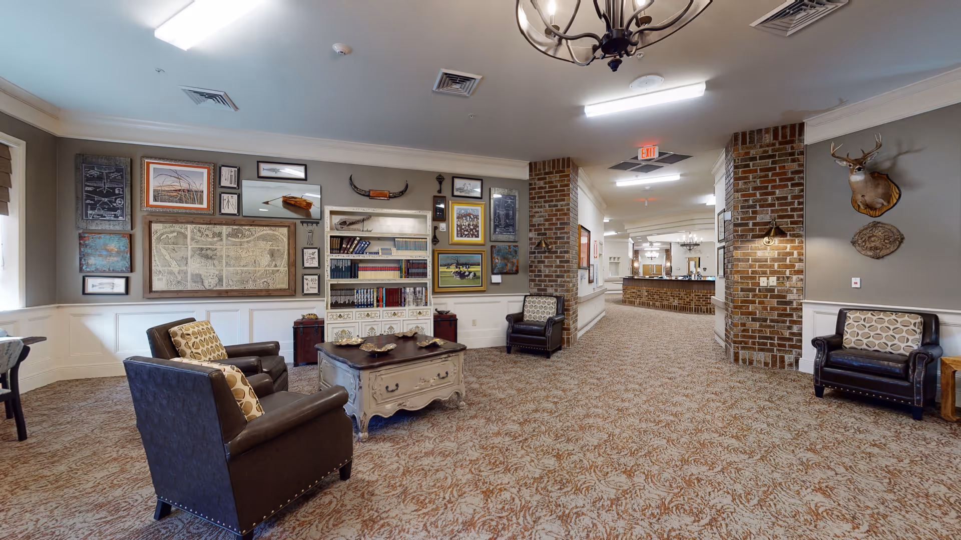 A cozy common area in a senior living facility featuring patterned carpet, several leather armchairs with patterned cushions, a white wooden coffee table, and a wall decorated with framed pictures and artwork. The room has brick pillars, a chandelier, and a mounted deer head on the wall. In the background, there is a hallway leading to another area with more seating and lighting fixtures.