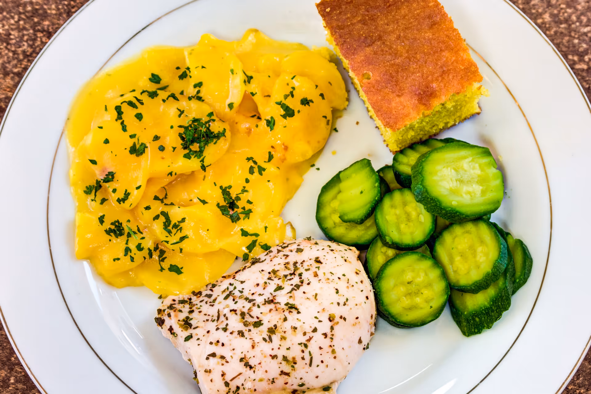 A white plate with a serving of scalloped potatoes garnished with herbs, a piece of seasoned cooked chicken breast, sliced cucumbers, and a square piece of cornbread.