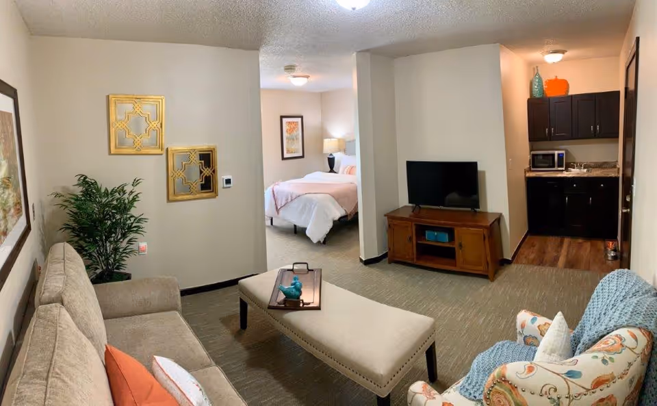 A cozy assisted living apartment featuring a living area with a beige sofa, an upholstered bench coffee table, and a patterned armchair with a blue throw blanket. The living room has decorative wall mirrors and a potted plant. In the background, there is a bedroom with a bed, nightstand, and lamp. To the right, a small kitchenette with dark cabinets, a microwave, and a sink is visible.