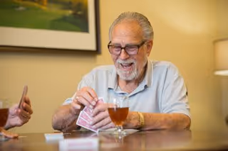 An elderly man with glasses and a beard is smiling and holding playing cards while sitting at a table with a glass of iced tea in front of him. Another person's hand is partially visible holding cards on the left side of the image. The setting appears to be a cozy indoor room with a framed picture on the wall and a lamp in the background.