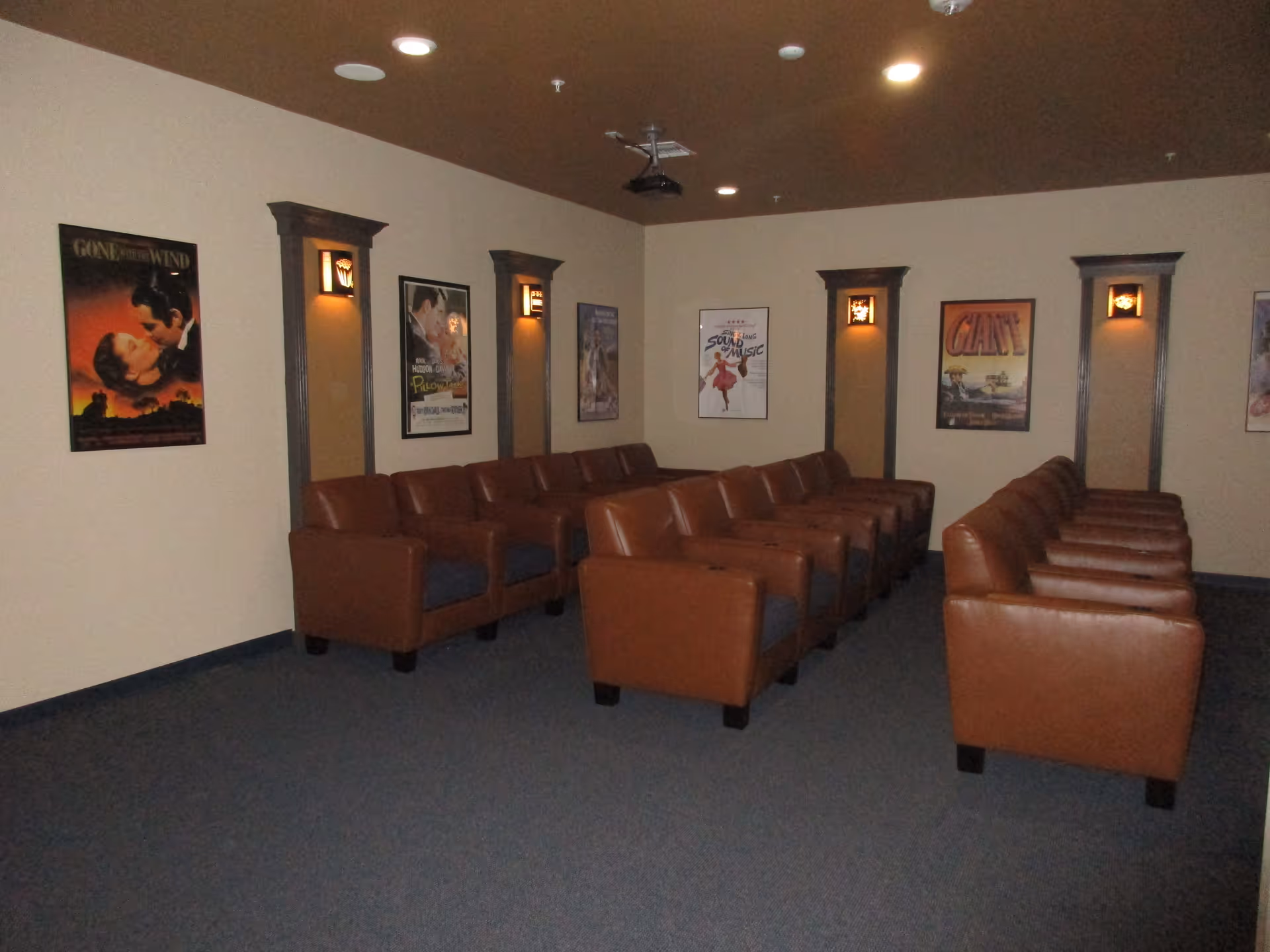 A small theater room with three rows of brown leather chairs facing a blank wall. The walls are decorated with framed classic movie posters including Gone with the Wind, The Sound of Music, and Giant. The room has dim wall sconces and a ceiling-mounted projector.