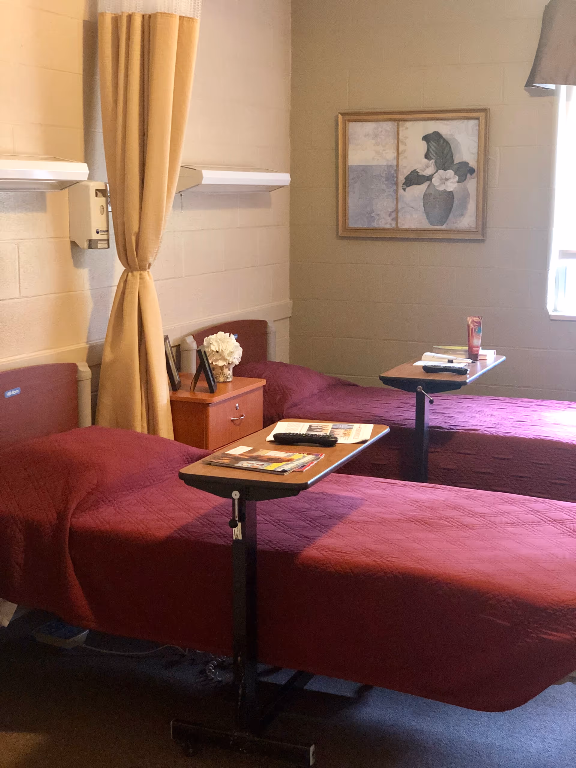 A room with two single beds covered in maroon bedspreads, each with an overbed table holding magazines and a remote control. Between the beds is a small wooden nightstand with a white flower arrangement and a framed photo. A beige curtain hangs on the wall behind the beds, and a framed painting of a vase with flowers is on the wall. A window with a beige valance lets in natural light.