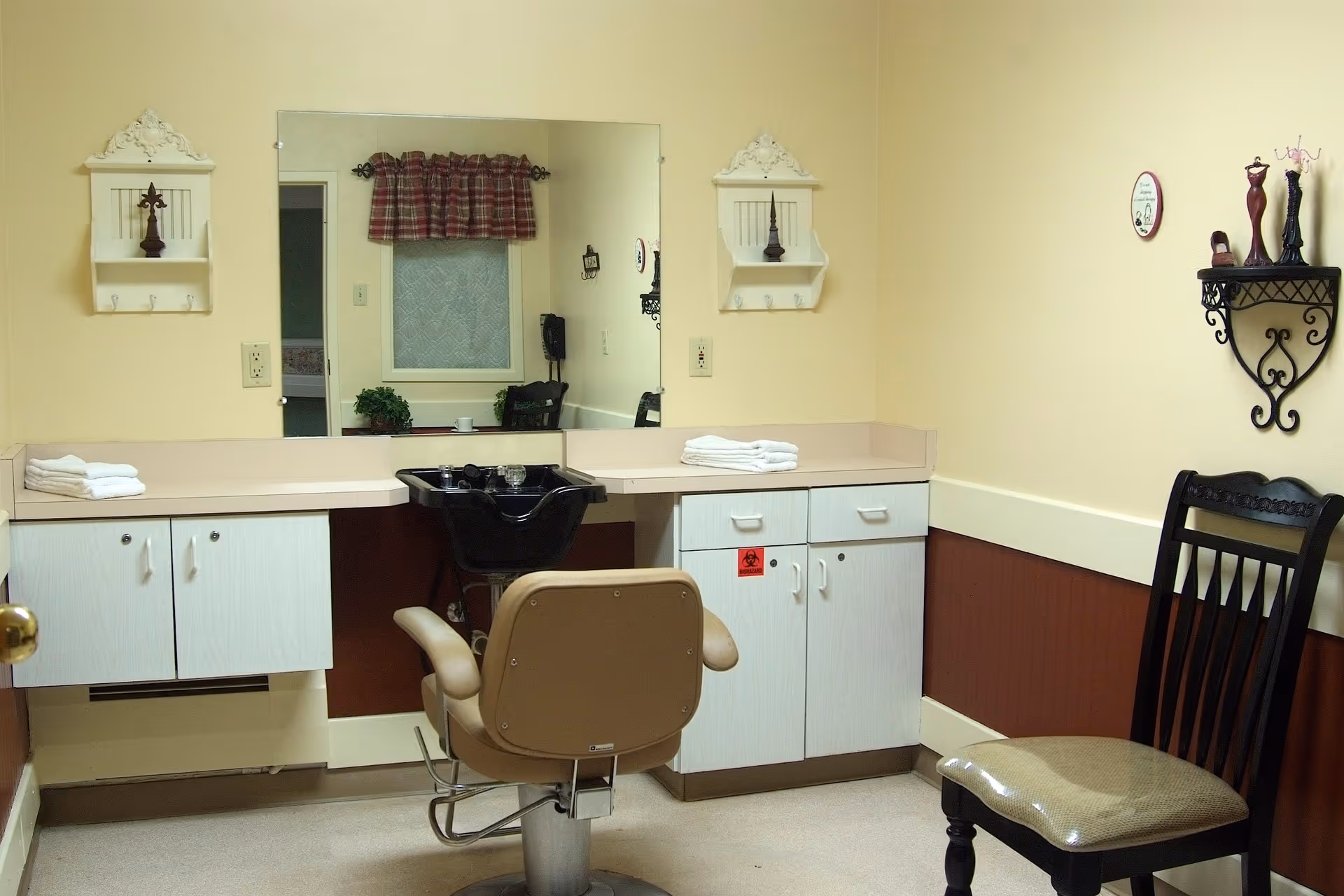 Interior of a small hair salon or grooming room with a beige salon chair facing a large mirror, a black wash basin, white cabinets with towels on the countertop, and a black wooden chair against the wall. The walls are painted yellow and brown with decorative shelves and small figurines.