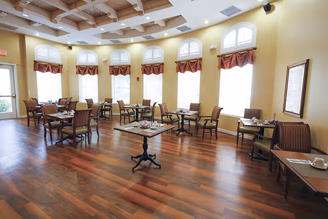A bright dining room with multiple small tables and chairs arranged neatly. The room has large arched windows with white shutters and red valances, wooden flooring, and a coffered ceiling with recessed lighting.