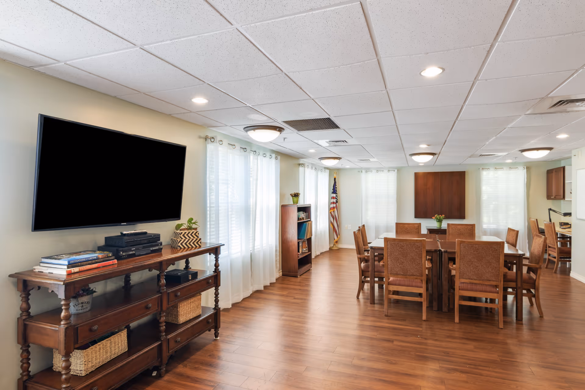 A spacious, well-lit common area with wooden flooring and beige walls. The room features a large flat-screen TV mounted on the wall above a wooden console table with books, baskets, and electronic devices. There are several wooden chairs arranged around a rectangular table in the center of the room. Large windows with sheer white curtains allow natural light to fill the space. An American flag stands in the corner near a bookshelf, and the ceiling has multiple recessed lights and fixtures.