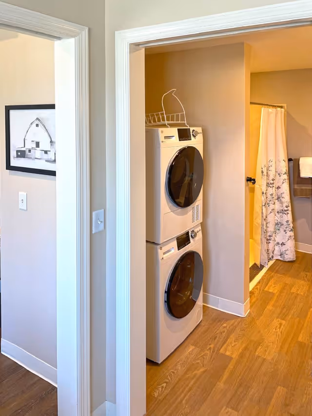 Interior view showing a stacked washer and dryer unit in a small laundry area next to a bathroom with a shower curtain and towel rack. The floor is wooden, and there is a framed black and white picture on the wall outside the laundry area.