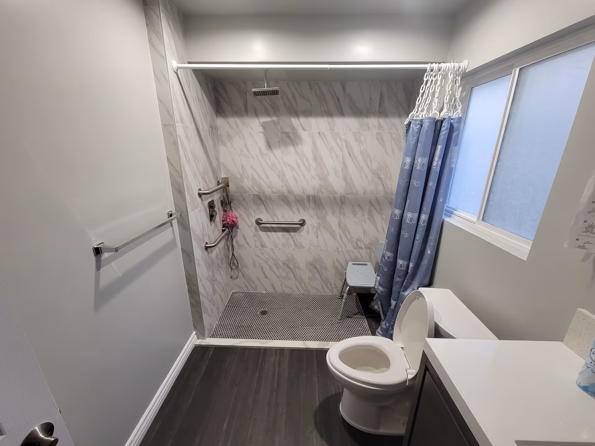 A modern bathroom with a walk-in shower featuring marble-patterned tiles and a rain shower head. The shower area has grab bars and a small shower chair. There is a blue shower curtain partially drawn to the side. The bathroom also includes a white toilet with the lid open, a towel rack on the wall, a frosted window, and a white countertop with a dark cabinet underneath.