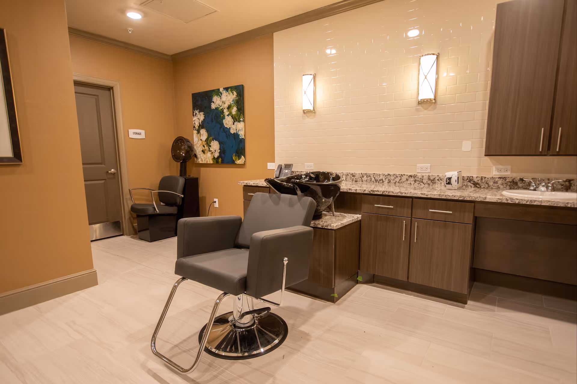 Interior view of a salon area in an assisted living facility featuring a gray salon chair in front of a black hair washing sink. The room has beige walls, a granite countertop with wooden cabinets, two wall-mounted lights, a painting with white flowers on a blue background, and a door labeled 'Storage'.
